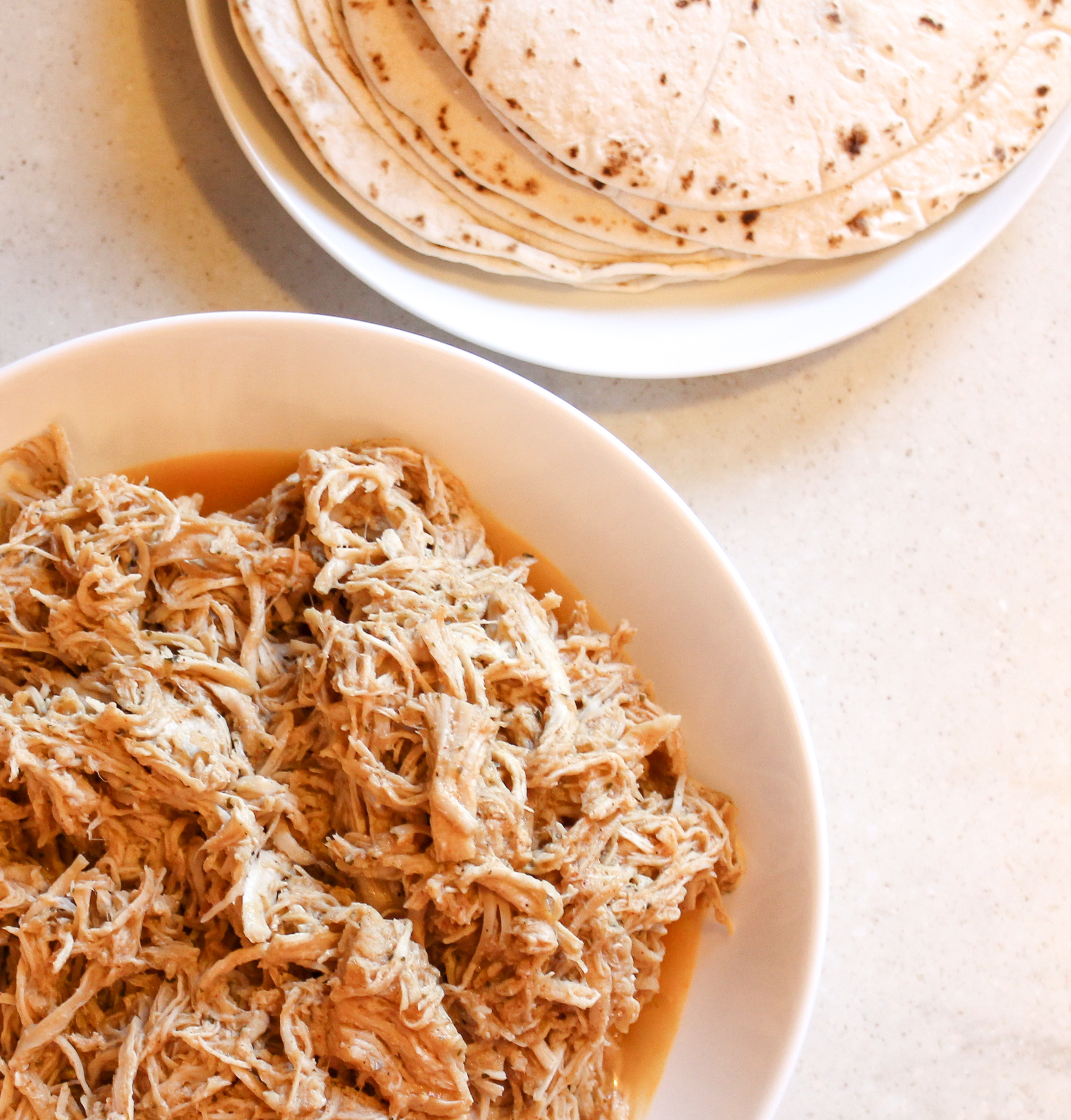 shredded chicken in a white bowl with a plate or tortillas beside the chicken