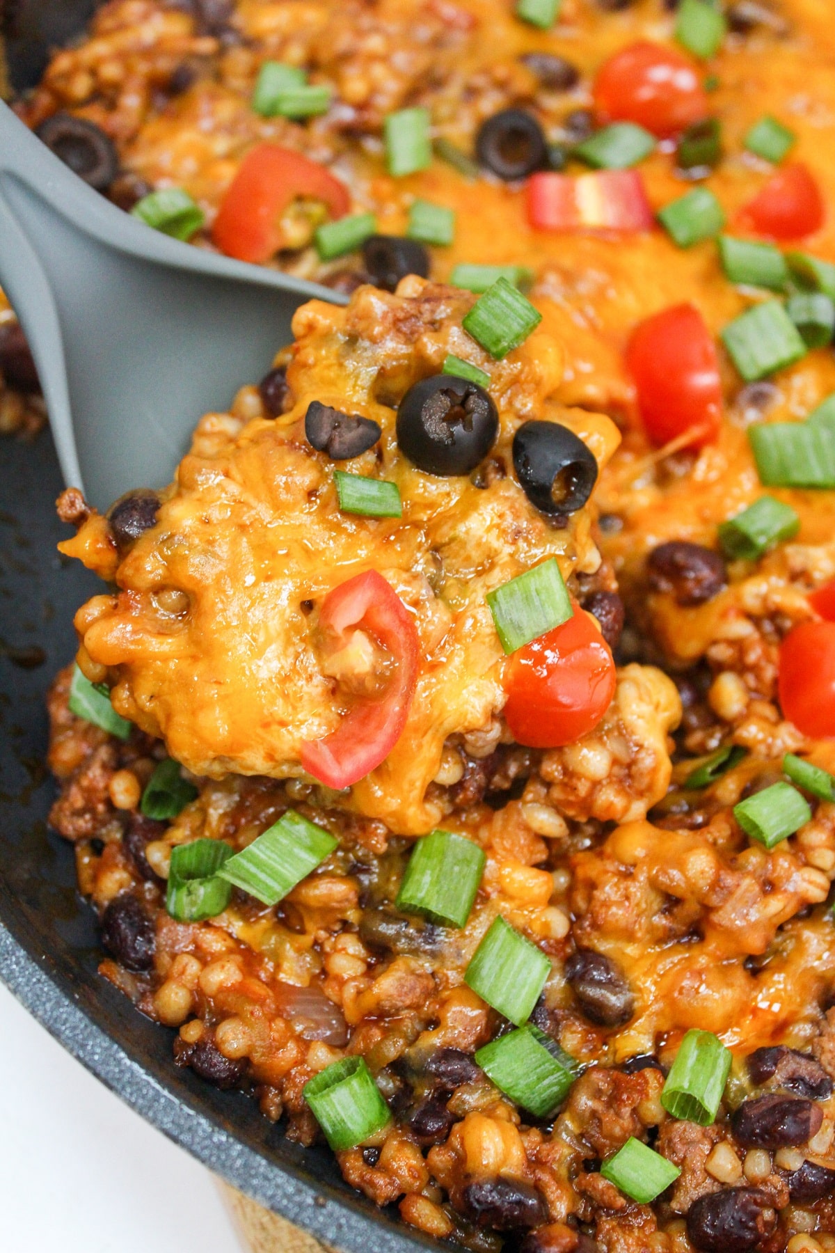 Cheesy Taco Barley Skillet cooked in a pan topped with cheese, black olives, tomatoes, and green onion. Scooped from the skillet with a serving spoon.