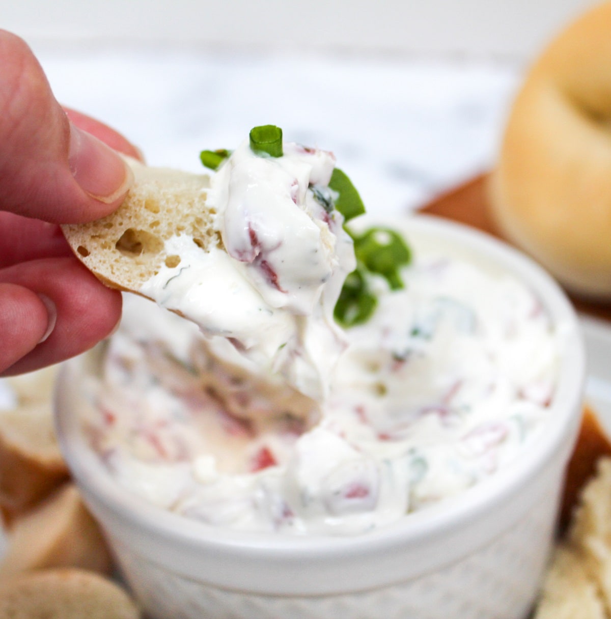 bagel dip in a white dish with bagel piece held in hand and dipped