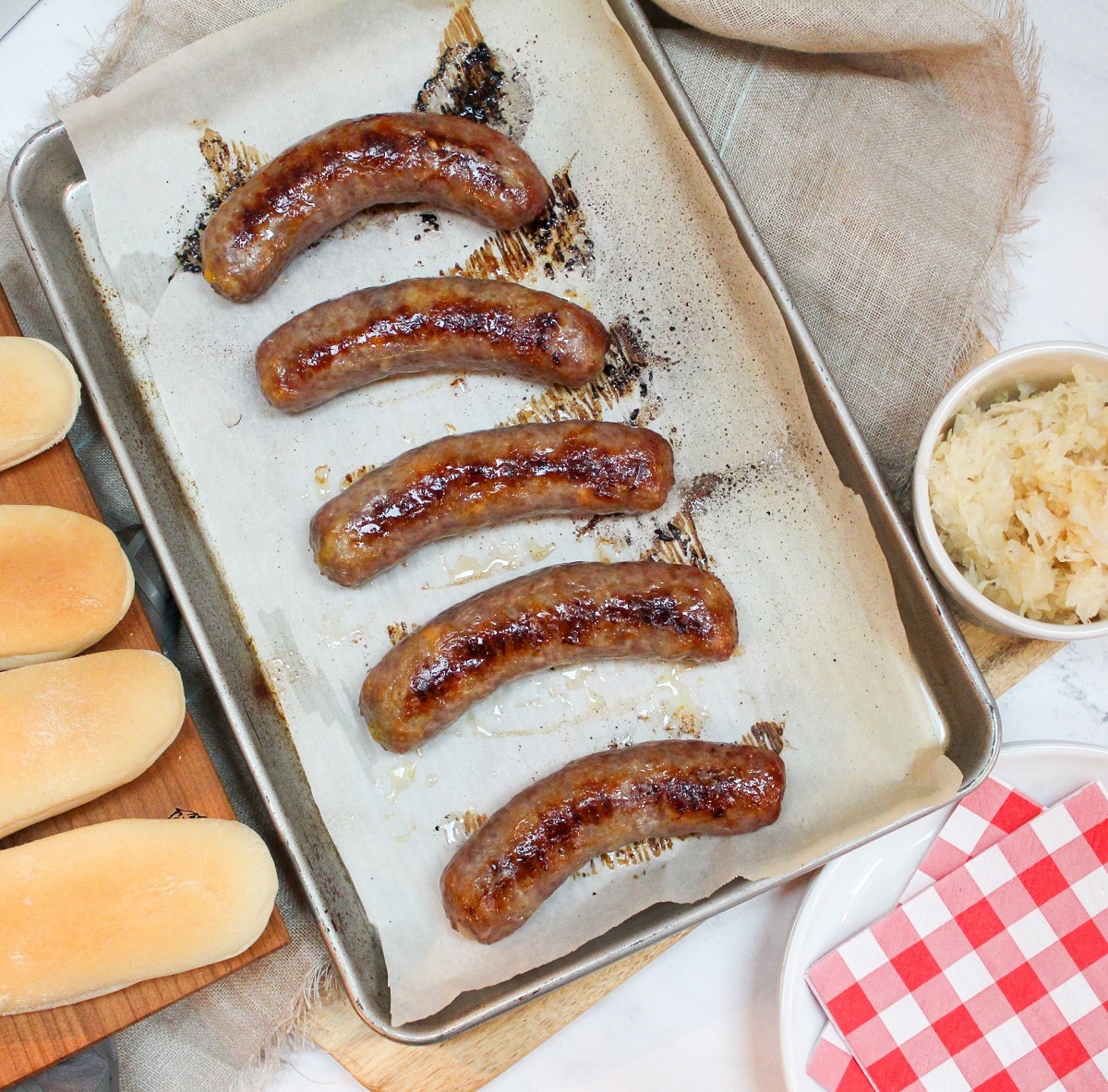 overhead photo of oven baked brats on a baking sheet with buns on the side and a bowl of sauerkraut.