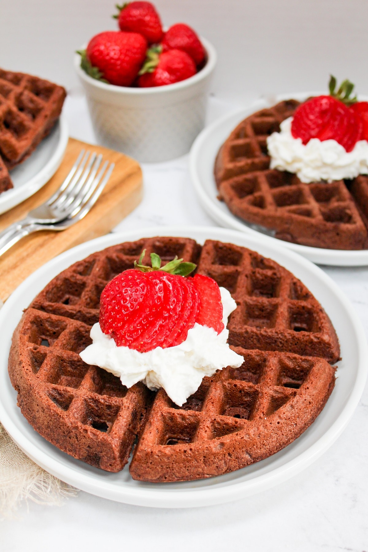 Brownie mix waffles with whipped cream and strawberries on top on a white plate with a plate of waffles in the background