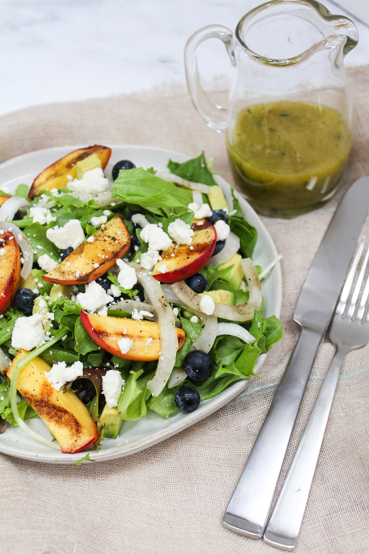 Lettuce topped with siced onions, grilled peaches, blueberries, and feta. There is a bowl of dressing and a fork on the side.
