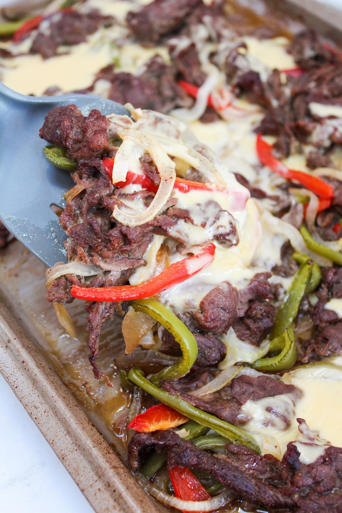 cooked sliced beer with cooked red and green peppers on a sheet pan. The sandwich filling is topped with a melted white cheese sauce. There is a spatula scooping the cheesesteak from the pan.