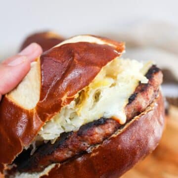 grilled brat patty on a pretzel roll. The meat is cooked and topped with melted cheese, mustard, and sauerkraut. The burger is show in a sideview because it is held in hand.