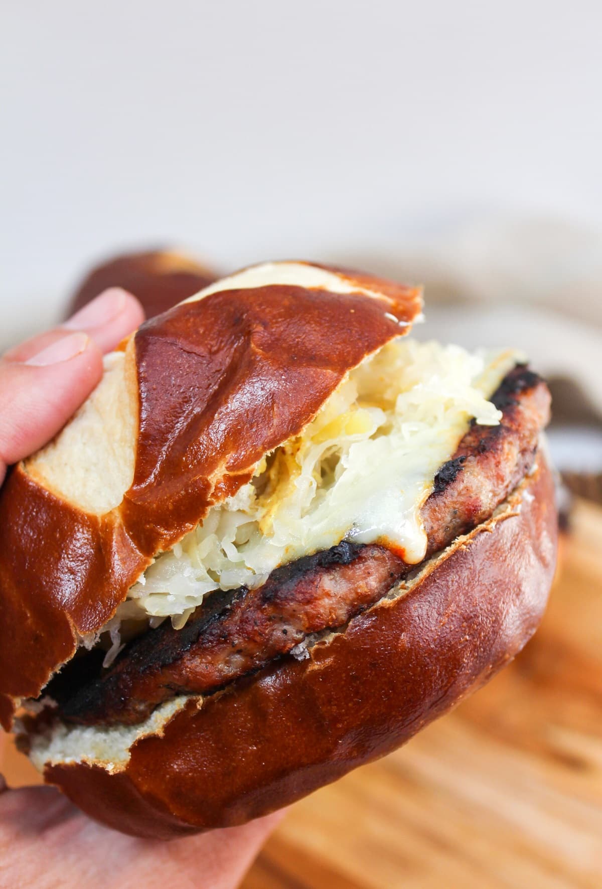 grilled brat pattie on a pretzel roll. The meat is cooked and topped with melted cheese, mustard, and sauerkraut. The burger is show in a sideview because it is held in hand.