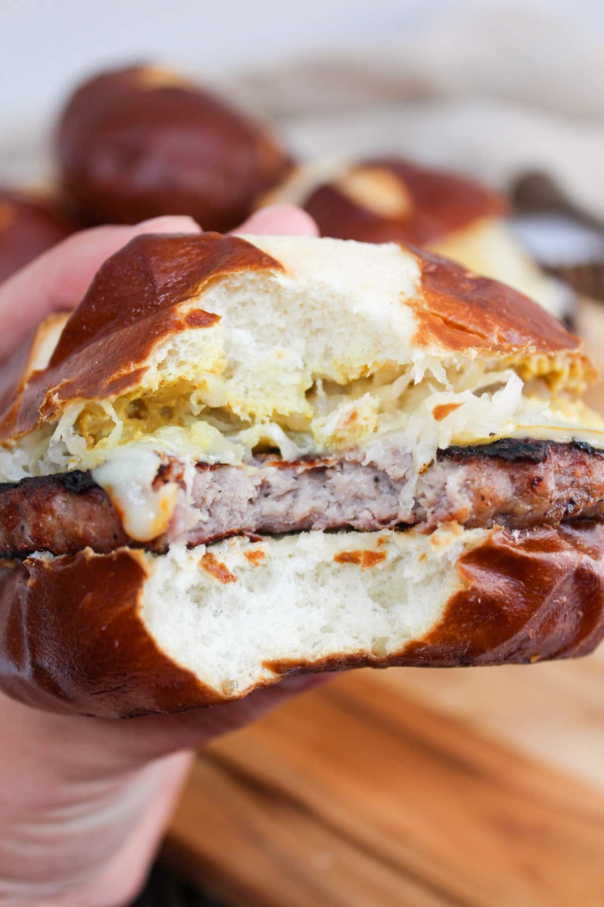 grilled brat pattie on a pretzel roll. The meat is cooked and topped with melted cheese, mustard, and sauerkraut. The burger is held in hand and there is a bite taken from the center.