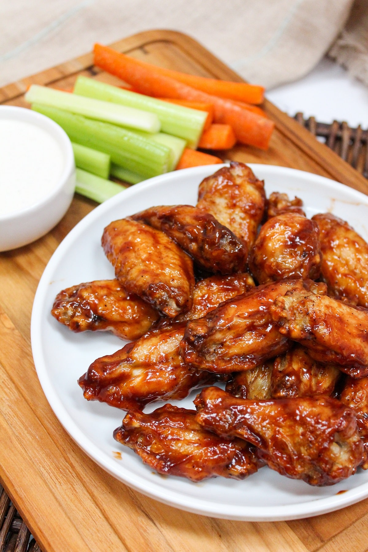 cooked wings in bbq sauce piled on a white plate. Sliced carrots and celery on the side with ranch dressing.