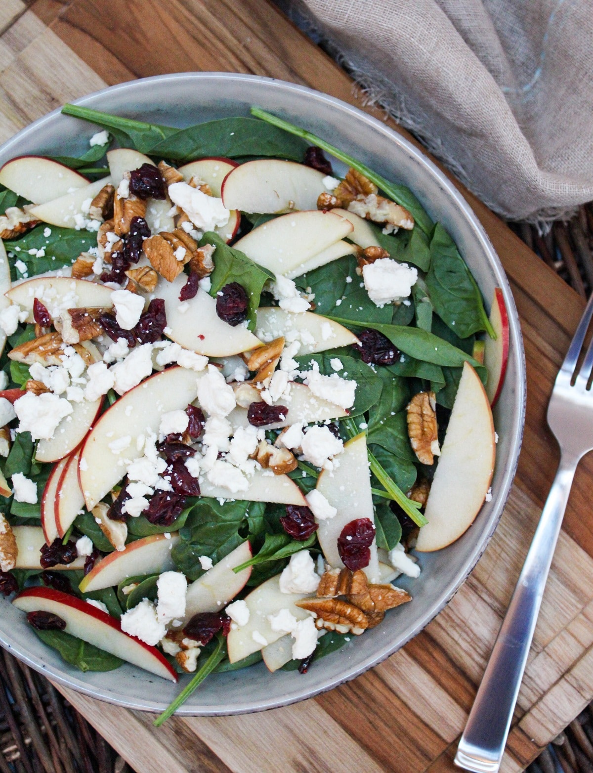 spinach in a gray bowl topped with sliced apples, pecans, crumbled feta cheese, and dried cranberries.