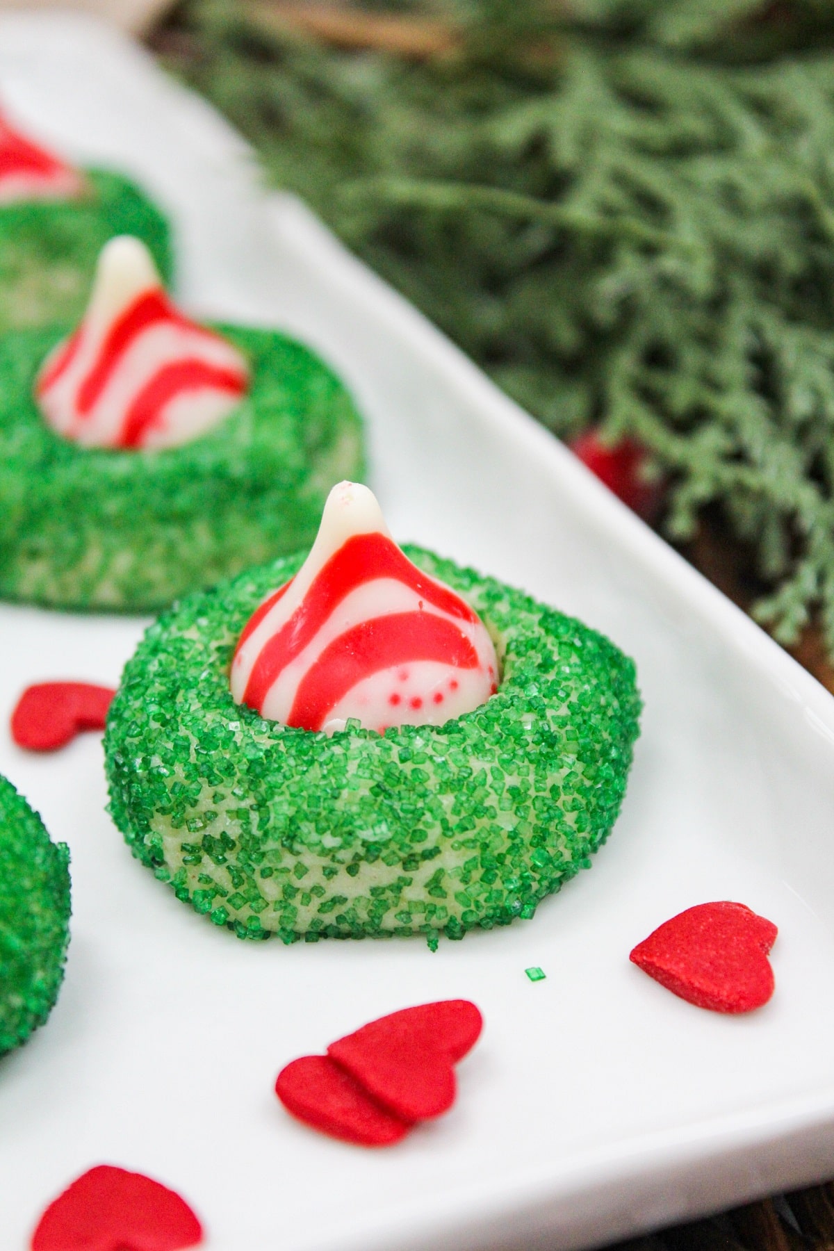 sugar cookies with small green sprinkles. A red and white kiss candy is placed in the middle. Cookies are served on a white plate. 