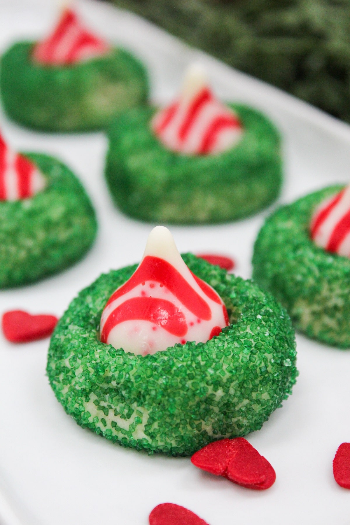 sugar cookies with small green sprinkles. A red and white kiss candy is placed in the middle. Cookies are served on a white plate.