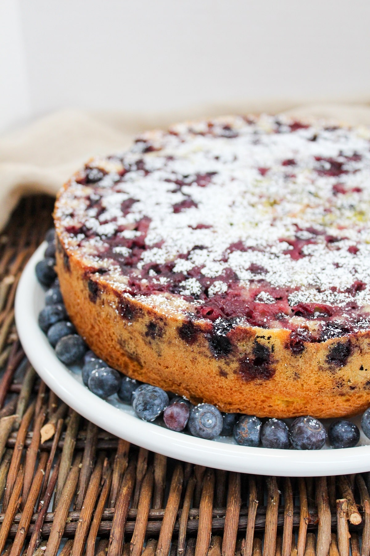 blueberry lemon cake on a white plate topped with powdered sugar and surrounded by fresh blueberries.