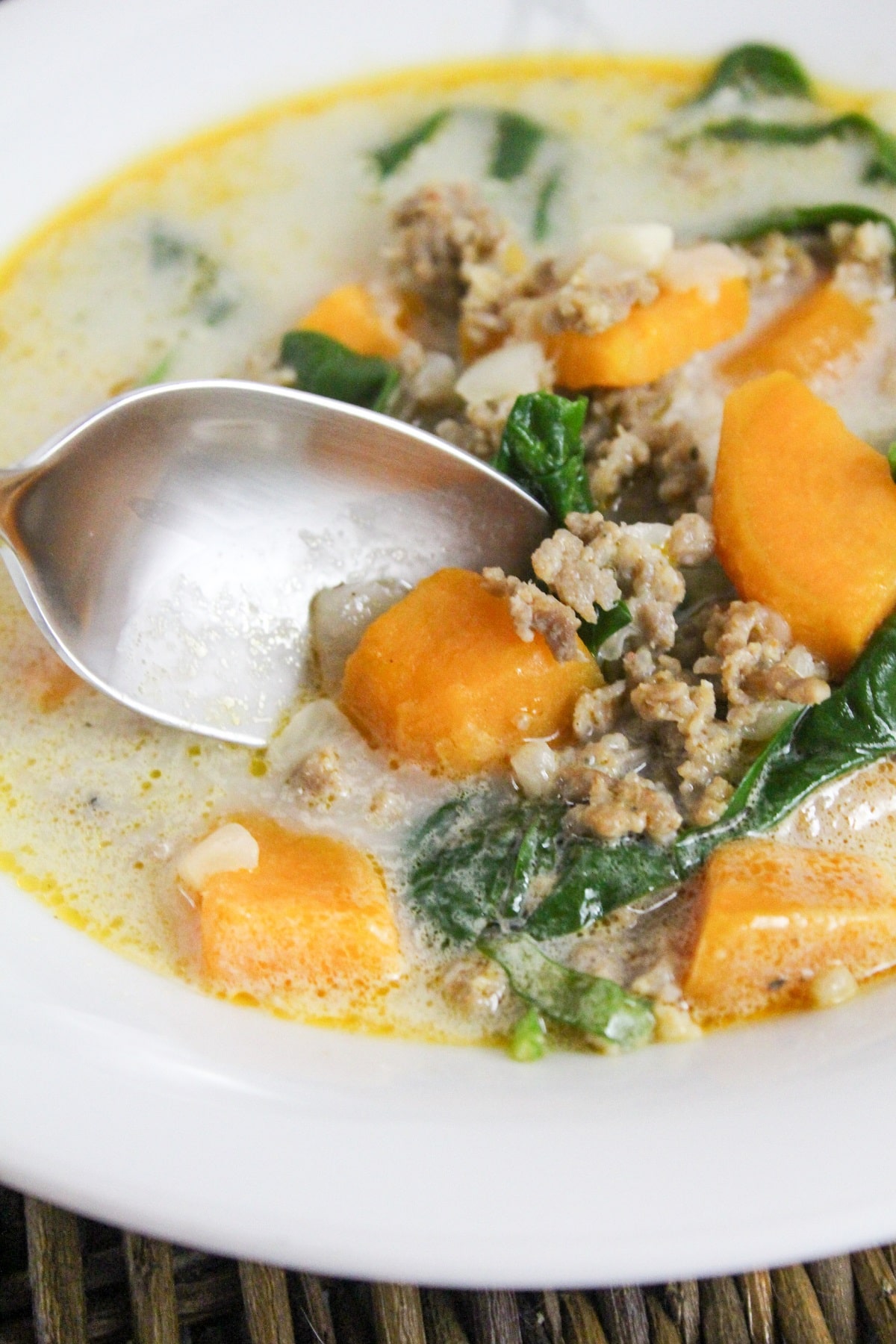 soup filled with sweet potatoes, ground sausage, and spinach with a spoon in a white bowl.