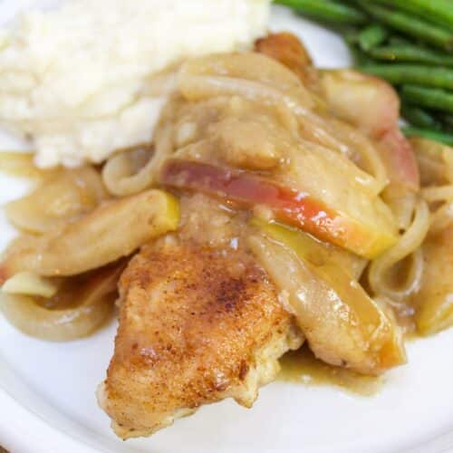 Cooked and browned chicken breasts on a white plate with apples and onions.