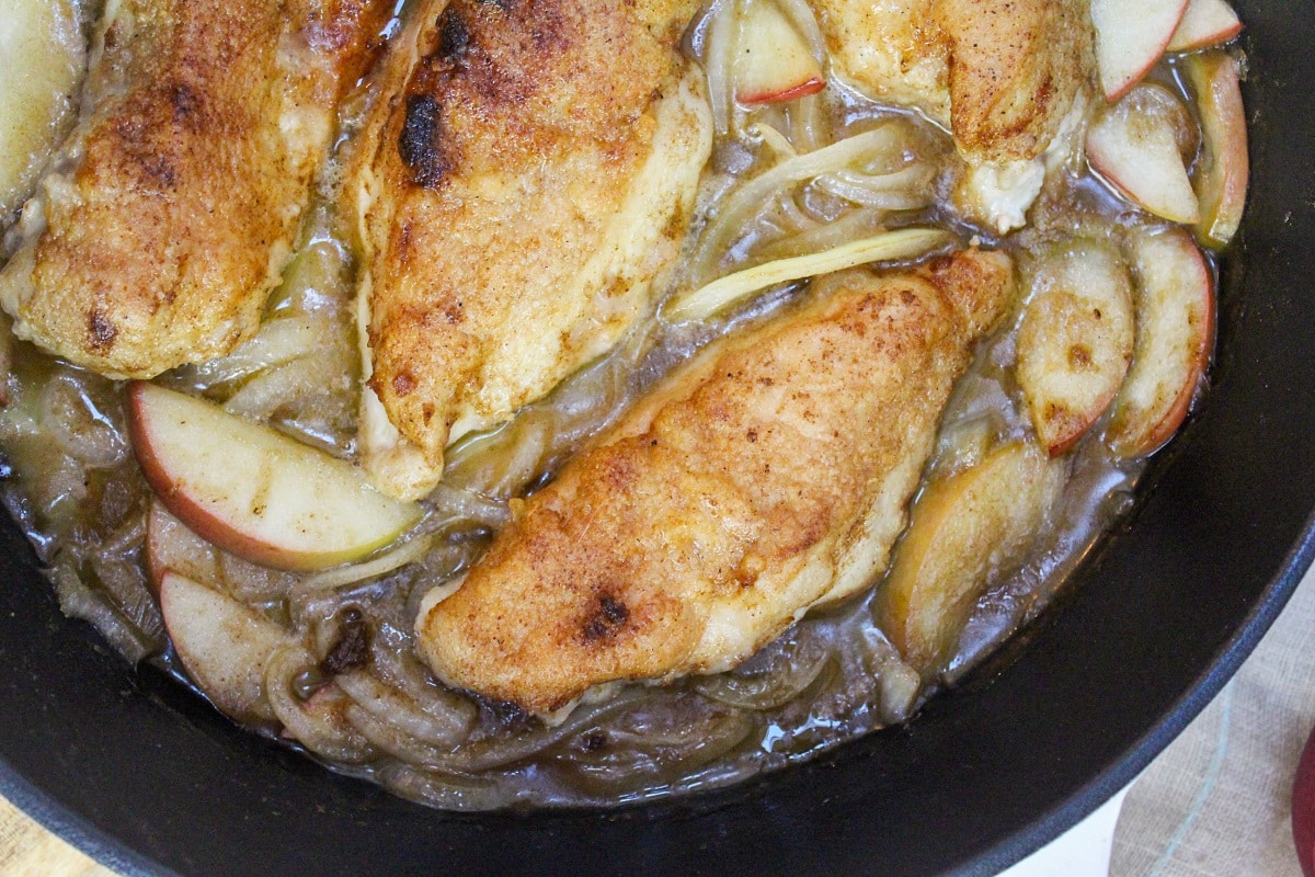 Cooked and browned chicken breasts in a black skillet with apples and onions.