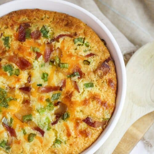 corn casserole topped with bacon, green onion, and cheese in a white baking dish.
