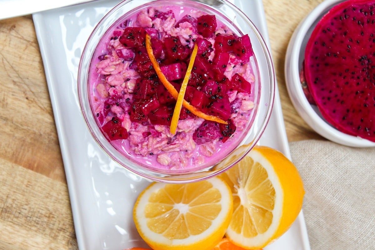 Pink overnight oats topped with chopped dragon fruit and citrus zest in a glass bowl.